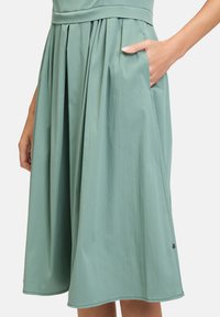Ein knielanger Rock aus zartem, mintgrünen Stoff mit Falten, einem taillierten Bund und seitlichen Taschen. Mit einem kleinen Logo am Saum.