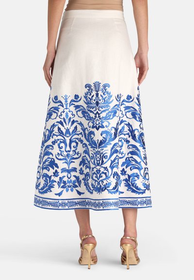 Žena v bielej midi sukni s prepracovanou modrou kvetinovou výšivkou, v kombinácii so zlatými sandálmi na vysokom opätku, fotografovaná zozadu.