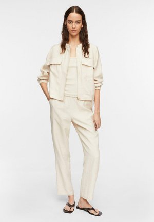 Kobieta z długimi brązowymi włosami, ubrana w jasnobeżową kurtkę, pasujące spodnie, top i czarne sandały z odkrytymi palcami, stojąca z rękami w kieszeniach.