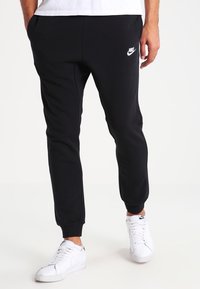 I pantaloni della tuta neri Nike presentano un design affusolato, una texture morbida, polsini elasticizzati e un accento di logo bianco sulla coscia sinistra. Indossati con scarpe da ginnastica bianche.