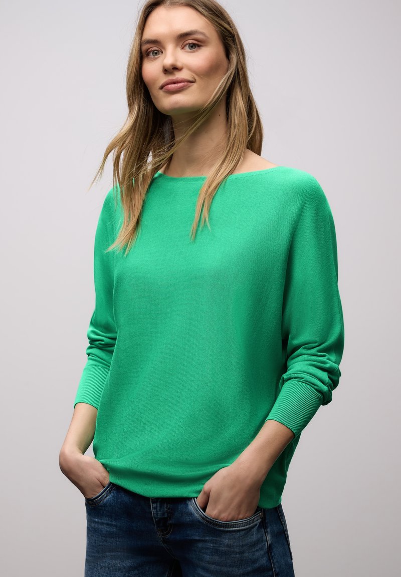 Femme aux cheveux longs portant un pull vert vif et un jean bleu, debout les mains dans les poches sur un fond clair uni.