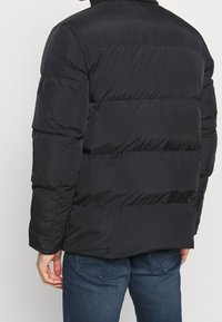 Giacca a vento nera imbottita con design trapuntato, caratterizzata da un colletto alto, una finitura testurizzata e cuciture a vista. Include una patch sulla manica sinistra.