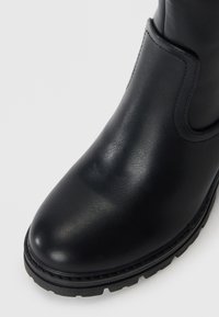 Botte chaussure en cuir noir avec une finition lisse, bout rond et panneau latéral élastique. Dispose d'une semelle en caoutchouc texturée avec un motif en relief.