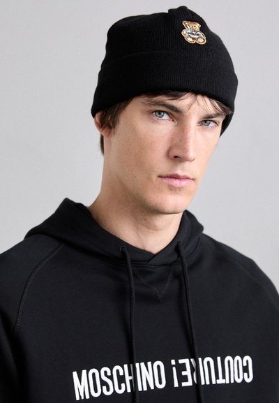 Schwarze Strickmütze mit einem Bärenmotiv in Gold und Braun. Getragen mit einem schwarzen Hoodie, auf dem "MOSCHINO COUTURE" in weißem Text steht.