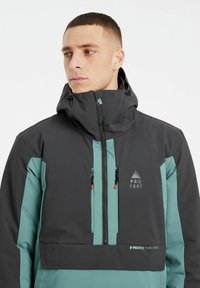 Sort og teal vandtæt jakke med høj krave, justerbar hætte, frontlynlås og to lynlåslommer. "PROTEST" logo vist.