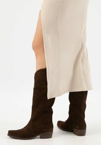 Brązowe zamszowe botki do kolan z lekko zaokrąglonym czubkiem i niskim obcasem, zestawione z beżową sukienką o prążkowanej fakturze z bocznym rozcięciem.