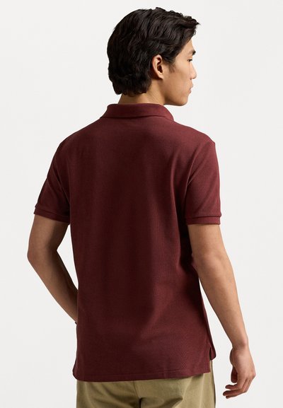 Polo shirt scurt maron cu un material texturat, guler și slituri laterale la tiv. Vederea din spate arată o siluetă ajustată.