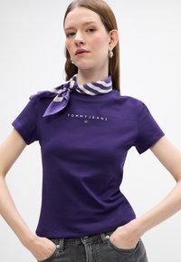 Violeta kokvilnas T-kreksa ar pieguļošu dizainu, uz kuras ir uzraksts "TOMMY JEANS" baltā krāsā, un gaismas krāsas svītrains šalle, kas sasieta ap kaklu.