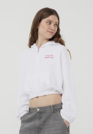 Felpa bianca corta con zip frontale, con testo ricamato rosso. Polsini e vita elasticizzati, abbinata a pantaloni in denim grigi.