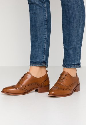 Bruine leren dresschoenen met een lage blokhak, voorzien van brogue-details en een ronde neus. Draagt met een spijkerbroek.
