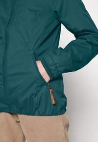 Zelenomodrá zipová bunda s žebrovanými manžetami a bočními kapsami. Na zipu je dřevěná úchytka a bunda má hladký, mírně lesklý povrch.