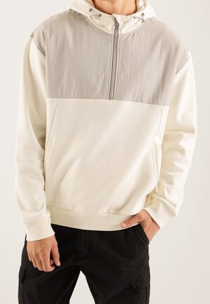 Homme portant un sweat à capuche crème et gris clair avec une fermeture éclair à l'avant, mains partiellement dans les poches, associé à un pantalon cargo noir.