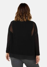 Zwarte top met lange doorschijnende mouwen, een solide lichaam en een iets langere achterkant. De textuur van het materiaal contrasteert met de gladde accenten op de schouders.