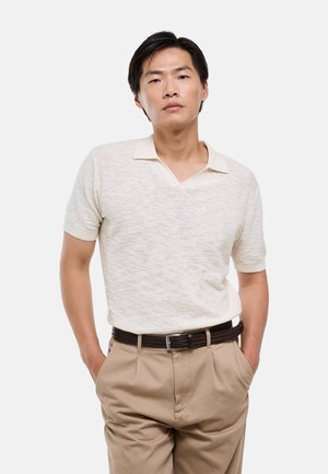 Homme aux cheveux courts et noirs porte une chemise crème à manches courtes avec col et un pantalon beige avec ceinture foncée, debout les mains dans les poches sur fond blanc.