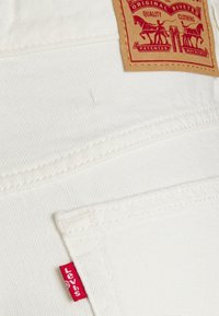 Tela de mezclilla blanca con costuras visibles y una etiqueta roja de "Levi's". Presenta una etiqueta marrón con el logotipo y el texto, cosida en la zona del bolsillo trasero.