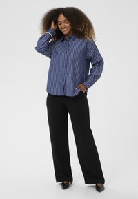 Zila un baltā svītraina krekla ar garām piedurknēm, kombinēts ar melnām platām biksēm un melniem ausītajiem apaviem, kas ir ar kabatām.