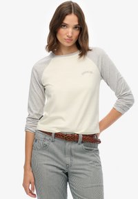 Vrouw draagt een cr èmekleurig en grijs shirt met lange mouwen met het "Superdry"-logo, gestreepte broek en een gevlochten bruine riem, poseert met één hand op haar heup.