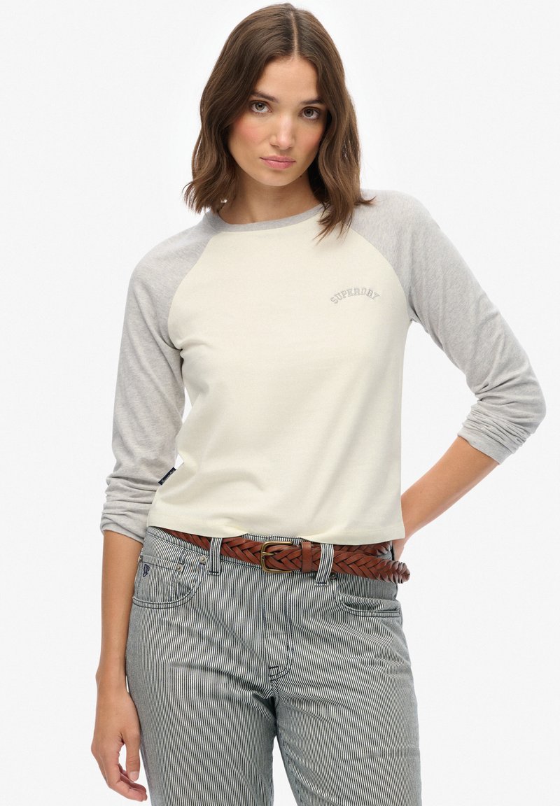 Vrouw draagt een crèmekleurig en grijs shirt met lange mouwen met het "Superdry"-logo, gestreepte broek en een gevlochten bruine riem, poseert met één hand op haar heup.