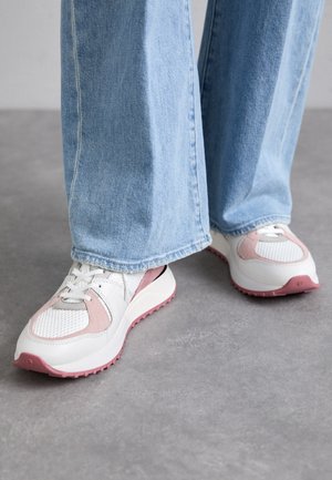 Des baskets blanches et roses dotées de matériaux en maille et en cuir, d'une semelle épaisse et d'accents texturés, partiellement obscurcies par un pantalon en denim bleu.