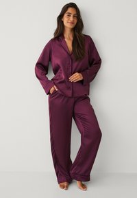 Satin pyjamasæt i dyb lilla, der har en knapfront skjorte med krave og lange ærmer samt matchende bukser med vide ben. Glat tekstur.