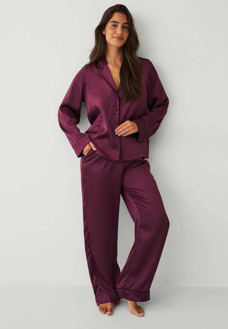 Satin pyjamasæt i dyb lilla, der har en knapfront skjorte med krave og lange ærmer samt matchende bukser med vide ben. Glat tekstur.