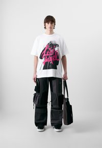 Witte oversized t-shirt met een roze grafische print, gecombineerd met zwarte cargobroek met meerdere zakken en zwarte schoenen.