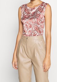 Blusa in satin con motivo paisley rosso e beige, abbinata a pantaloni beige in pelle plissettati. Primo piano sulla parte superiore del corpo.
