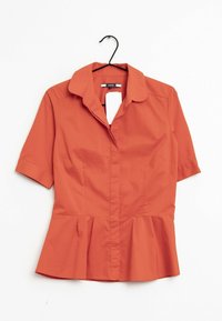 Blusa naranja de manga corta con cuello, frontal de botones, dobladillo peplum y textura de tela suave. Cuelga de un gancho negro sobre un fondo blanco.
