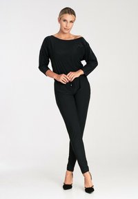 Czarny kombinezon z dekoltem na jedno ramię, rękawami o długości trzech kwart, dopasowanym pasem i zwężanymi nogawkami. Miękka, gładka faktura.