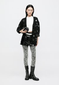 Cardigan negru cu model cu stele, top alb scurt, pantaloni scurți negri strălucitori, colanți cu imprimeu de șarpe și cizme negre până la genunchi. Ține o cameră foto în mâini.