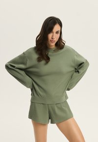 Jasnozielony sweter z prążkowanego materiału o luźnym kroju i szerokich rękawach, w zestawie z dopasowanymi krótkimi szortami, obie części mają miękką teksturę.