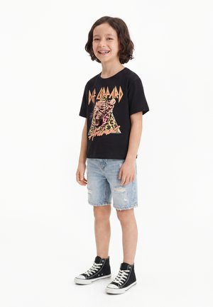 Enfant souriant aux cheveux bouclés portant un t-shirt noir du groupe Def Leppard, un short en jean clair déchiré et des baskets montantes noires, debout sur un fond blanc.