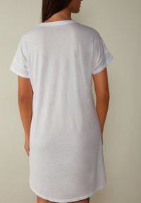 Abito in cotone bianco a t-shirt con maniche corte arrotolate e scollatura rotonda. Texture morbida, lunghezza fino a metà coscia, senza motivi o accentuazioni visibili.