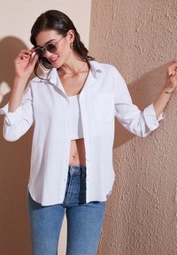 White, lightweight, long-sleeve shirt with a pocket, button-down front, and rolled cuffs, worn over a cropped top and paired with blue jeans.