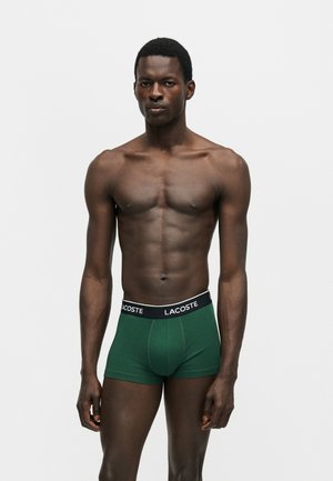 Homme à la peau foncée torse nu, portant un boxer vert Lacoste, debout devant un fond uni gris clair.