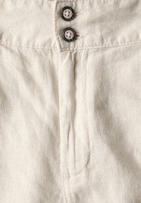 Linnenbyxor i en ljus beige färg, med två runda knappar, kontrastsömmar och en enda framficka med en subtil textur.