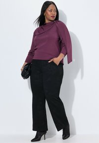 Bordový dlouhý rukáv s jemně draped detaily na rameni, v kombinaci s černými kalhotami s decentním vzorem. Modelka drží černou tašku a má na sobě boty na podpatku.