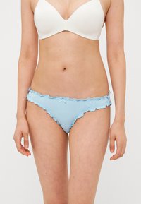 Vrouw in witte beha en lichtblauwe rimpelige bikini-onderbroek, staand tegen een effen achtergrond.