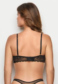 Frau mit braunem Haar, die einen schwarzen Spitzen-BH und passende durchsichtige Unterwäsche trägt, von hinten vor einem weißen Hintergrund gezeigt.