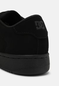 Zapatilla de ante negra con una suela de goma texturizada negra, que presenta un diseño cosido y un acento de logo negro en el talón.