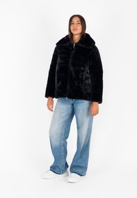 Giacca in pelliccia sintetica nera con ampio colletto e chiusura con zip, caratterizzata da una texture morbida. Abbinata a jeans a gamba larga di un leggero blu e scarpe bianche.