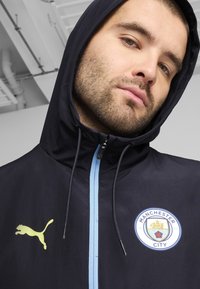 Hoodie nero con zip blu, con un logo Puma giallo sul petto sinistro e uno stemma rotondo del Manchester City sul davanti.