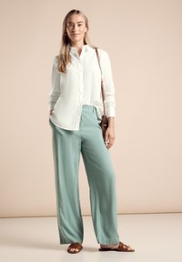 Camisa blanca de botones, pantalones anchos verde claro, sandalias marrones y un bolso cruzado de mimbre. Tejido suave y corte relajado.