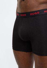Melnas bokseršorti, izgatavoti no gluda auduma, ar melnu jostiņu, kurā ir sarkans "HUGO" logotips. Pieguļošs siluets ar moderno dizainu.
