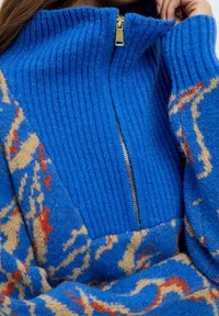 Niebieski wełniany sweter o prążkowanej fakturze, z półzamkiem przy kołnierzu i abstrakcyjnym wzorem w beżowych i czerwonych tonach. Charakteryzuje się miękkim wykończeniem.