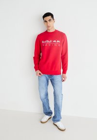Sudadera roja con un logo de "Red Bull Racing" en el pecho. Combinada con jeans de mezclilla azul claro y zapatillas blancas. Textura suave, ajuste relajado.