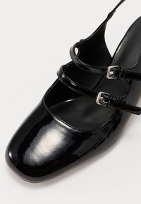 Chaussure en cuir verni noir avec un bout rond, comportant deux sangles à boucle sur le pied et une texture lisse.