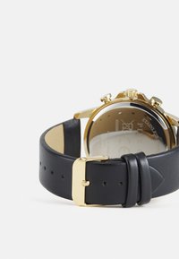 Montre-bracelet en cuir noir avec un boîtier en acier inoxydable doré, grande couronne et fonctions chronographe. Le dos en verre transparent met en valeur le mouvement.
