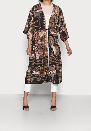 Femme portant une longue veste kimono noire à motif floral avec une ceinture à la taille, un pantalon blanc et des talons ouverts noirs.