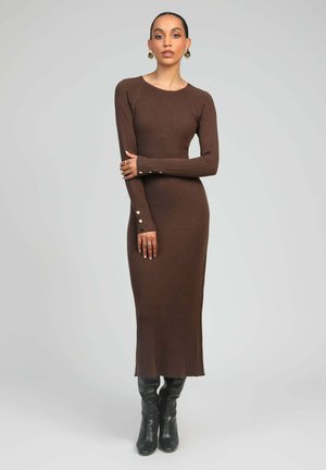 Robe longue à manches longues en tricot côtelé marron avec un col rond et des boutons en accent aux poignets. Portée avec des bottes noires montantes jusqu'aux genoux.